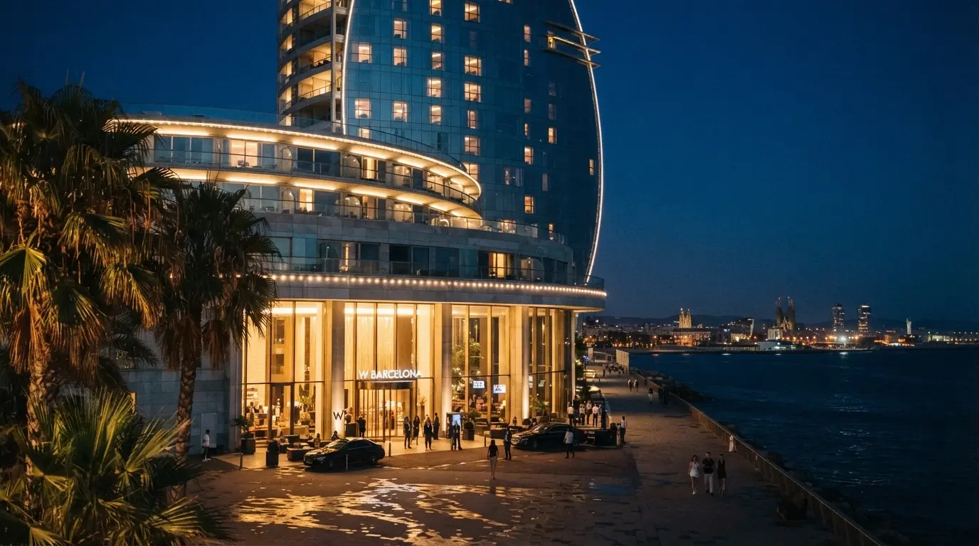 Fachada nocturna iluminada del Casino Barcelona en Port Olímpic, con la entrada principal visible, el mar al fondo y el skyline de Barcelona reflejado en los ventanales